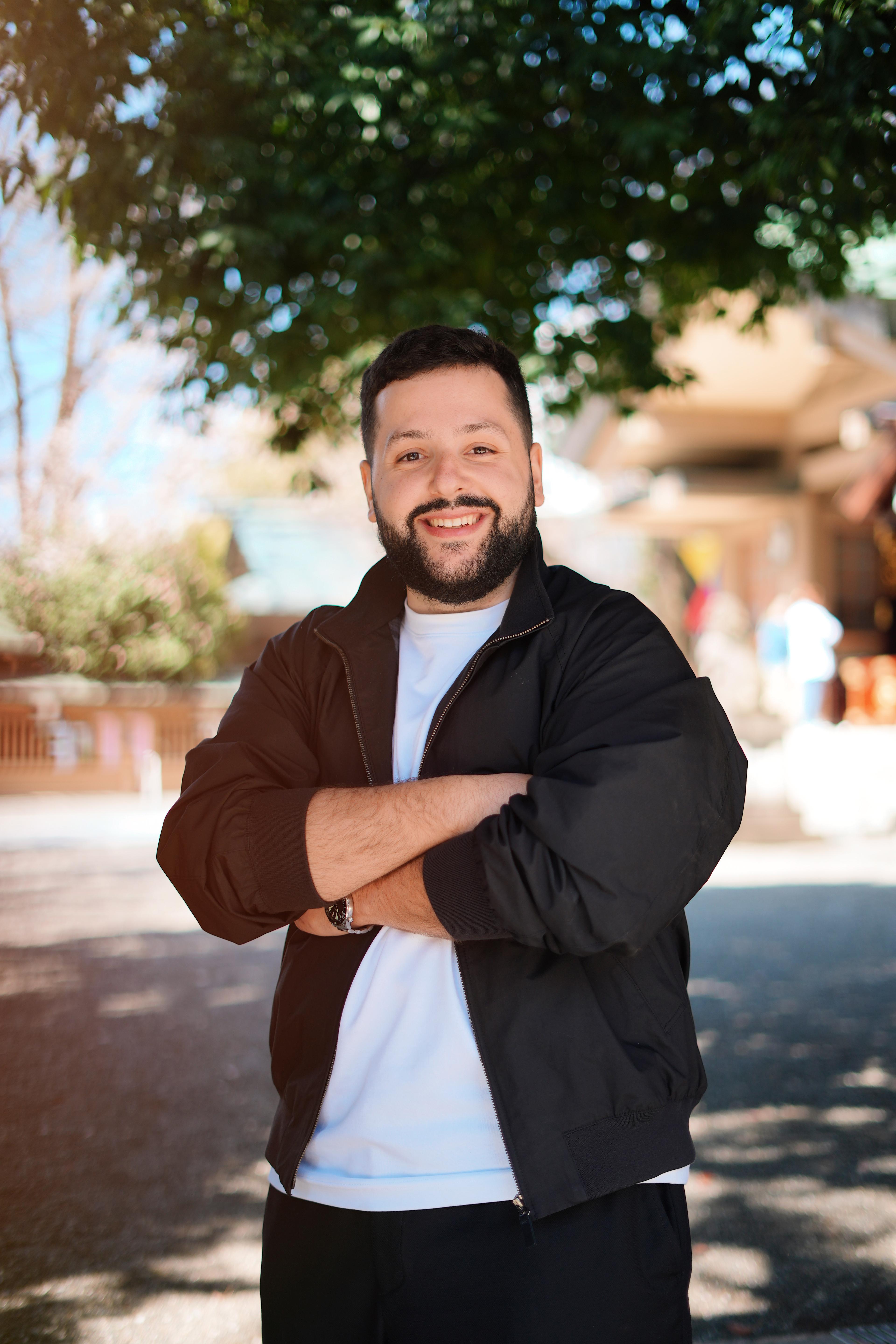 Founder portrait of Oriol: warm and approachable, wearing a black jacket over a white tee, in natural daylight against a soft-blurred urban street background.